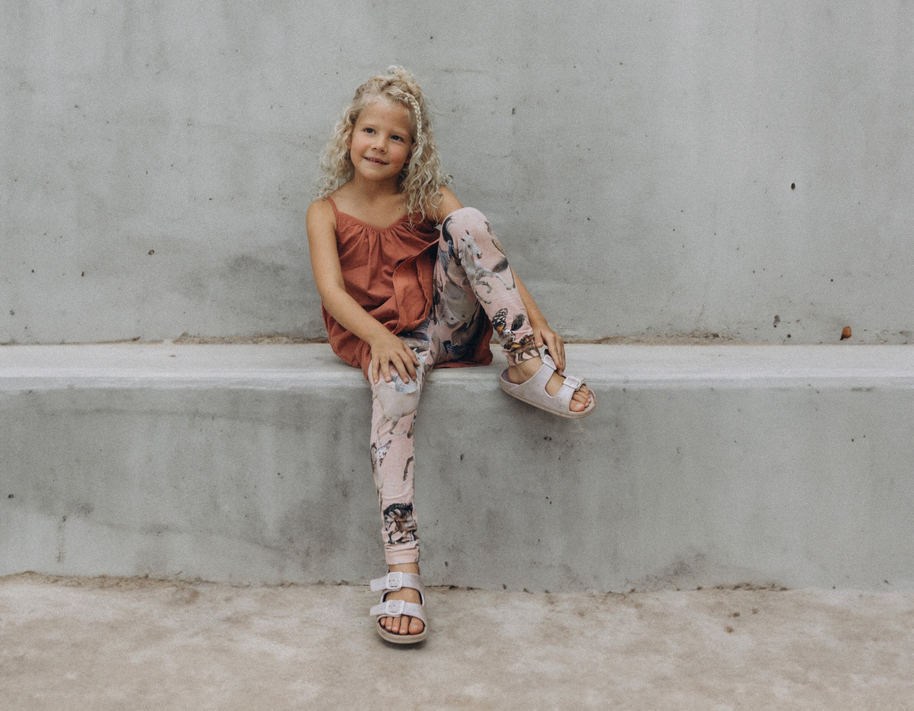 Little girl wearing Adaline dress and horse themed leggings, while sitting on a concrete bench.