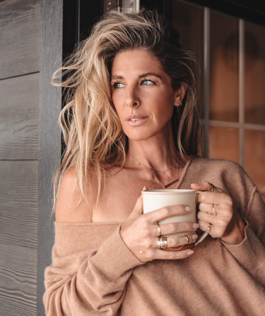 Angi leaning against farmhouse wall drinking coffee looking into the horizon.