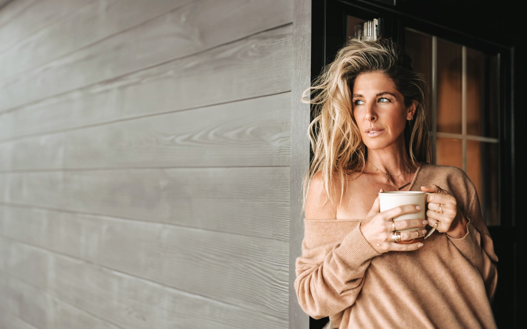 Angi leaning against farmhouse wall drinking coffee looking into the horizon.