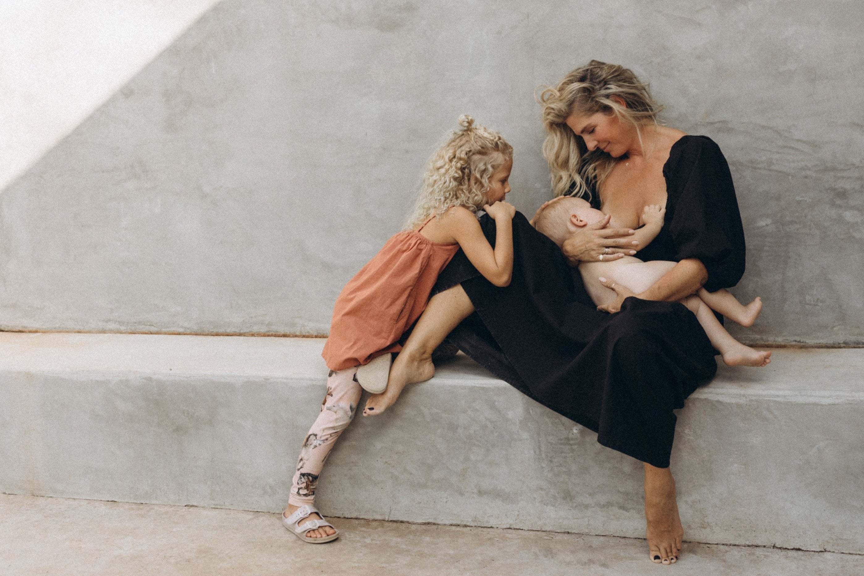 Angi breastfeeding in Adeline dress in Midnight Black color, with daughter looking on wearing Mamoosh Childrens dress, while sitting on a concrete bench.