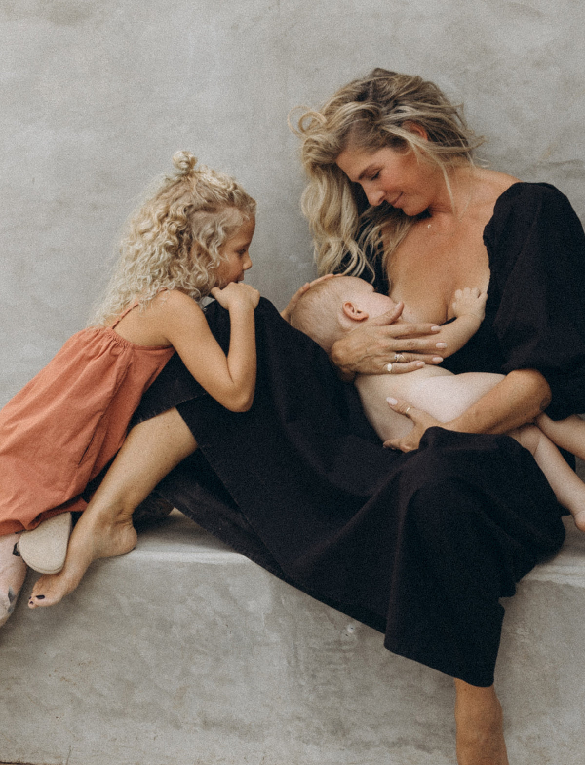 Angi breastfeeding in Adeline dress in Midnight Black color, with daughter looking on wearing Mamoosh Childrens dress, while sitting on a concrete bench.